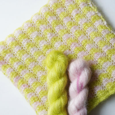 Two skeins of yarn, one green and one pink, on a textured yellow and white fabric background.