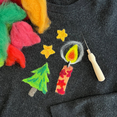 A close-up of a dark gray felted wool sweater decorated with colorful felted wool Christmas motifs, including a green tree, a red and yellow candle, yellow stars, and fuzzy wool accents. A needlepoint tool is resting on the chest.