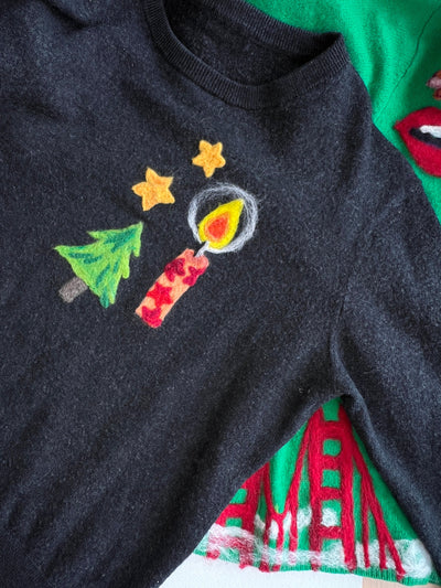 A close-up of a dark charcoal gray knit sweater featuring a hand-painted Christmas design with a green tree, red candle, and yellow stars, layered over a green sweater with red and white patterns.