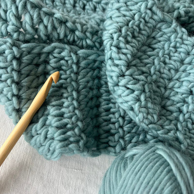 Teal knitted fabric with a crochet hook and yarn ball on a light gray background