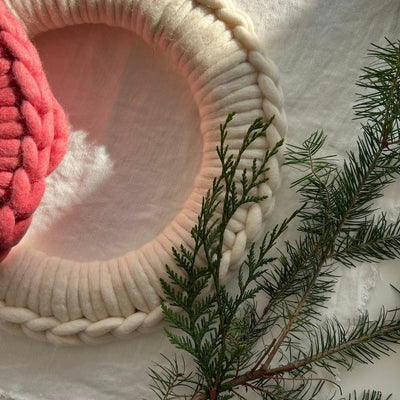 A chunky off-white crocheted wreath frame decorated with green pine branches, styled on a white fabric background with a pink knit item partially visible on the left.