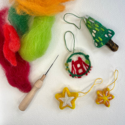 A flat-lay of handmade felt Christmas ornaments and wool roving on a white background, including a green tree ornament, a round ornament with a red bridge motif, two yellow flower/star ornaments, a wooden-handled needle tool, and colorful wool fibers in orange, green, red, and yellow.