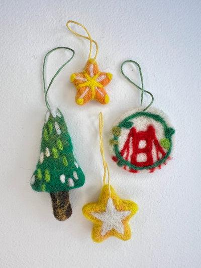 Four handcrafted felt Christmas ornaments on a white background: a green tree with white dots and brown trunk, a yellow-orange star, a round ornament with a red bridge motif and green border, and a yellow star with a white center.