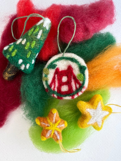 A set of handcrafted felted wool Christmas ornaments on a white background with colorful felted wool fibers behind them. The ornaments include a green felted Christmas tree with white accents, a round white ornament with red monogram lettering, and two star-shaped ornaments in yellow and orange.
