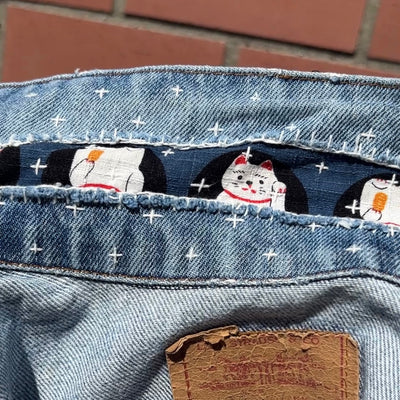Close-up of the inner collar of a light blue denim jeans jacket, showing a decorative pattern of white waving cats on a dark blue background with white star-like crosses made from sashiko embroidery stitches.
