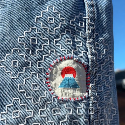 Close-up of a light blue denim garment with white geometric sashiko embroidery and a circular patch featuring a red sun and blue mountain motif.