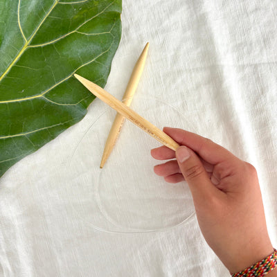 Hand holding a wooden knitting needle next to a green leaf on a white fabric background