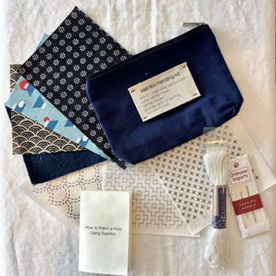 Sashiko mending kit with fabric pieces, navy pouch, and sewing supplies on a white background