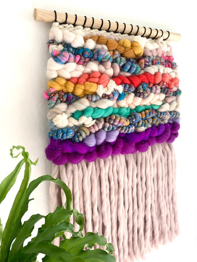 Colorful woven wall hanging with pom-poms and a plant in the foreground