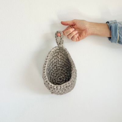A hand holding a small gray crocheted hanging basket with a loop handle, shown against a plain white wall.