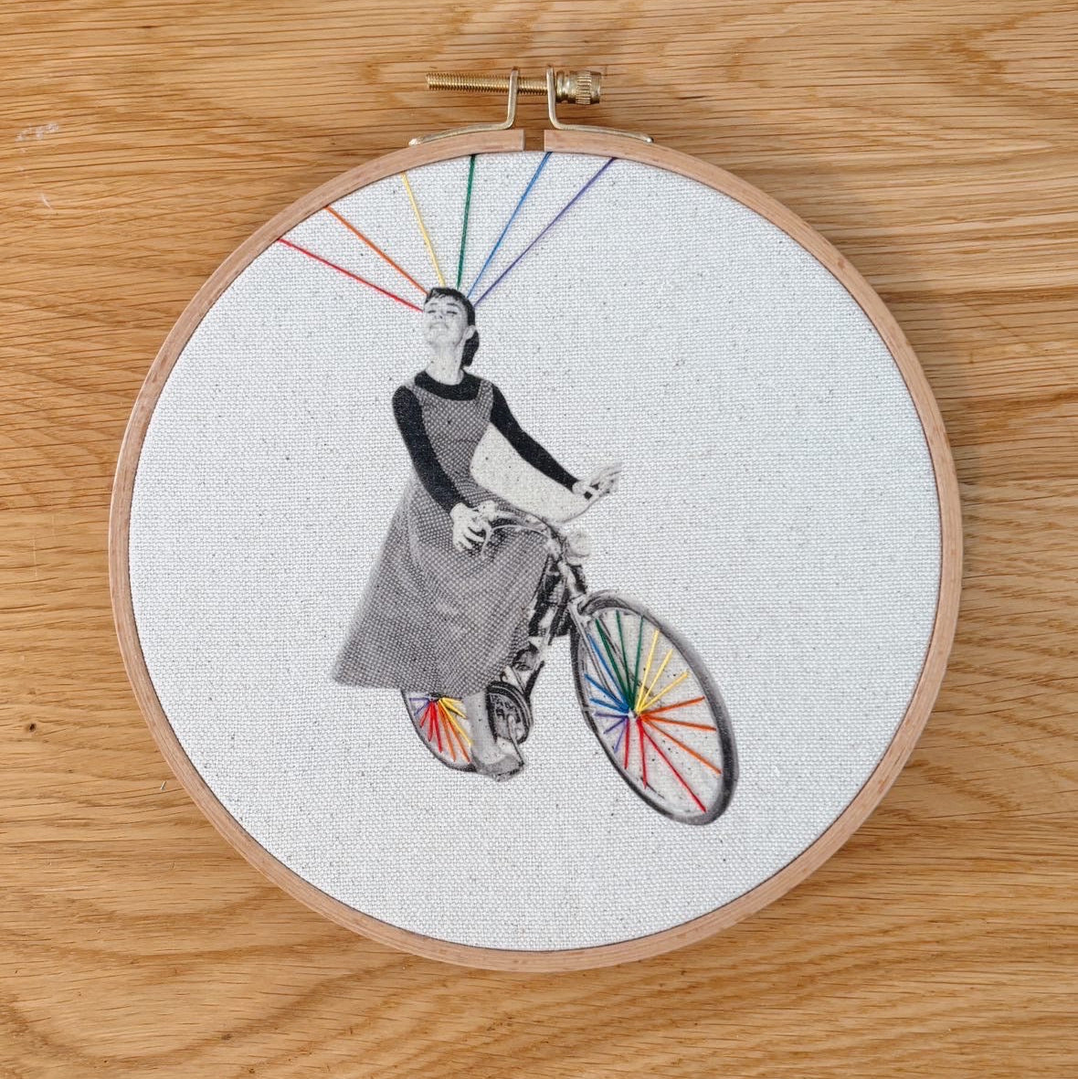 Embroidery hoop with a person riding a bicycle featuring rainbow wheels on a wooden surface