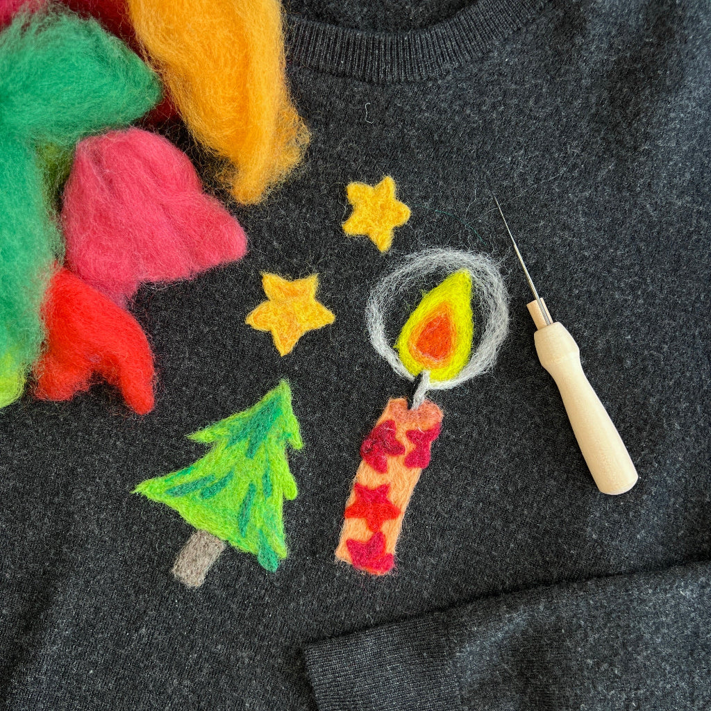 A close-up of a dark gray felted wool sweater decorated with colorful felted wool Christmas motifs, including a green tree, a red and yellow candle, yellow stars, and fuzzy wool accents. A needlepoint tool is resting on the chest.