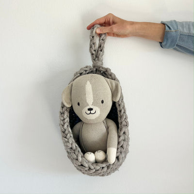 A gray knit dog plush toy sitting inside a chunky gray knit hanging basket, held by a person's hand against a plain white wall.