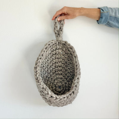 A hand holding a chunky gray crocheted hanging basket with a loop handle, set against a plain white wall.