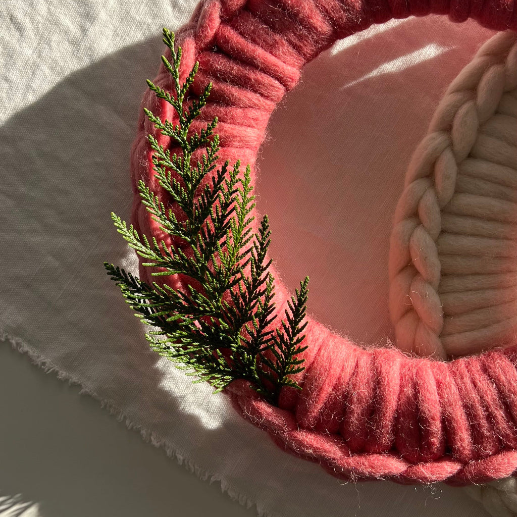 A handmade circular wreath made from chunky pink braided yarn, decorated with a small green pine sprig, resting on a textured white fabric surface under direct sunlight.
