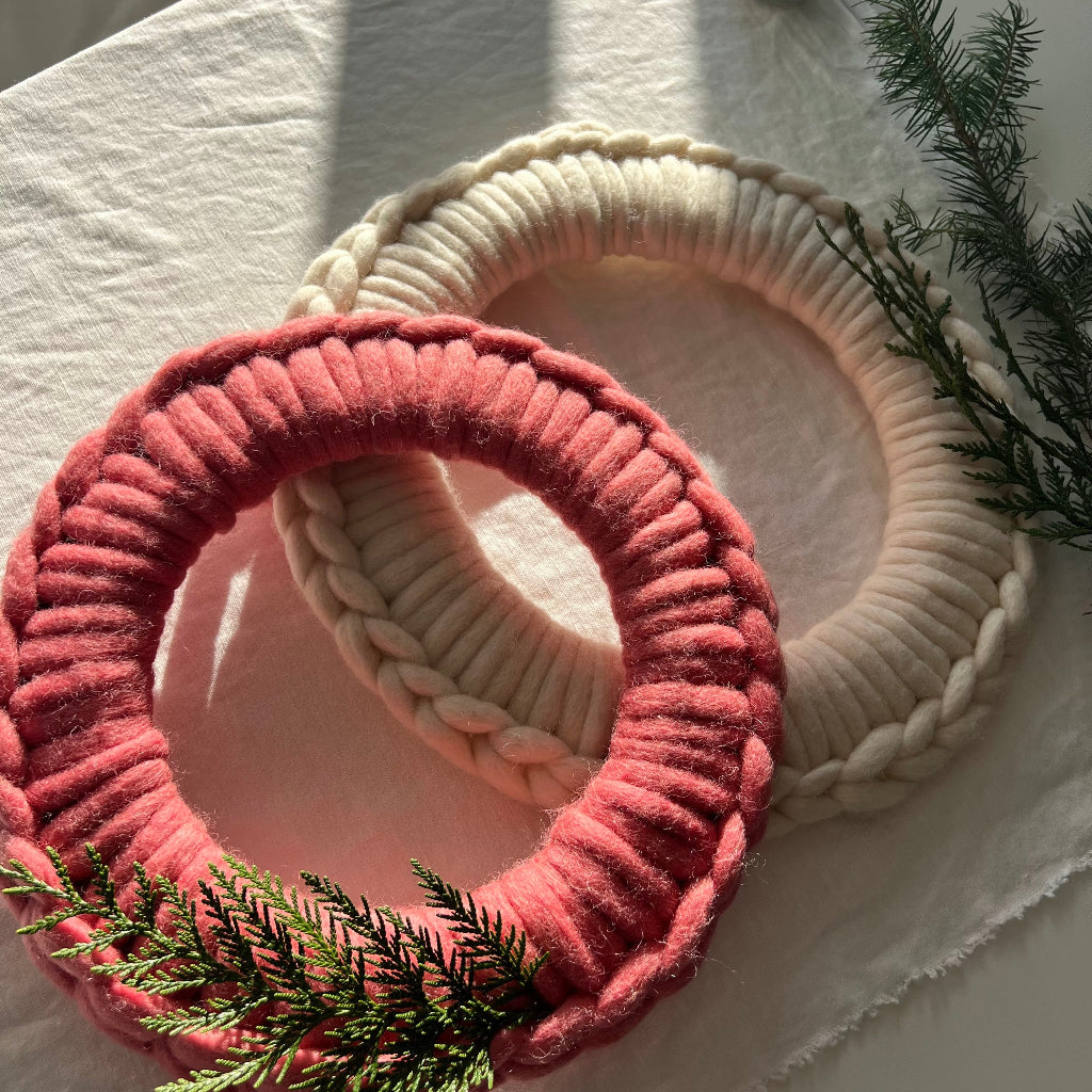 Two circular crocheted wool wreaths, one in dusty coral pink and one in cream, arranged on a white fabric background with green pine branches as styling props.