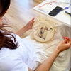 Person holding a round embroidery hoop with a design on a bed