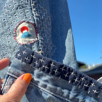 Close-up of a light blue denim garment with a decorative embroidered patch and a dark navy hem featuring white stitched cross details, held by a hand with orange nail polish against a blue sky background.
