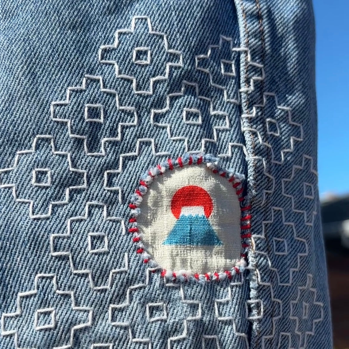 Close-up of a light blue denim garment with white geometric sashiko embroidery and a circular patch featuring a red sun and blue mountain motif.