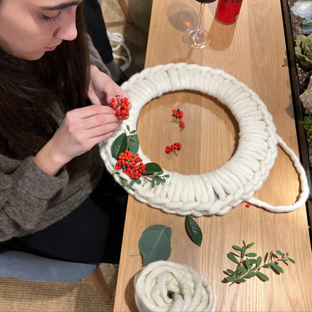 Person crafting a decorative wreath with white yarn and red berries on a wooden table.