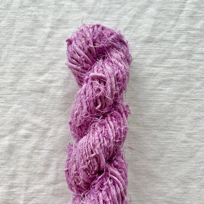 A skein of fuzzy, fringed yarn in a dusty lavender-purple color, laid vertically on a textured off-white linen background.