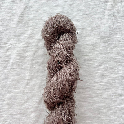A skein of fluffy, curly yarn in a muted taupe-brown color, laid vertically on a textured off-white fabric background.