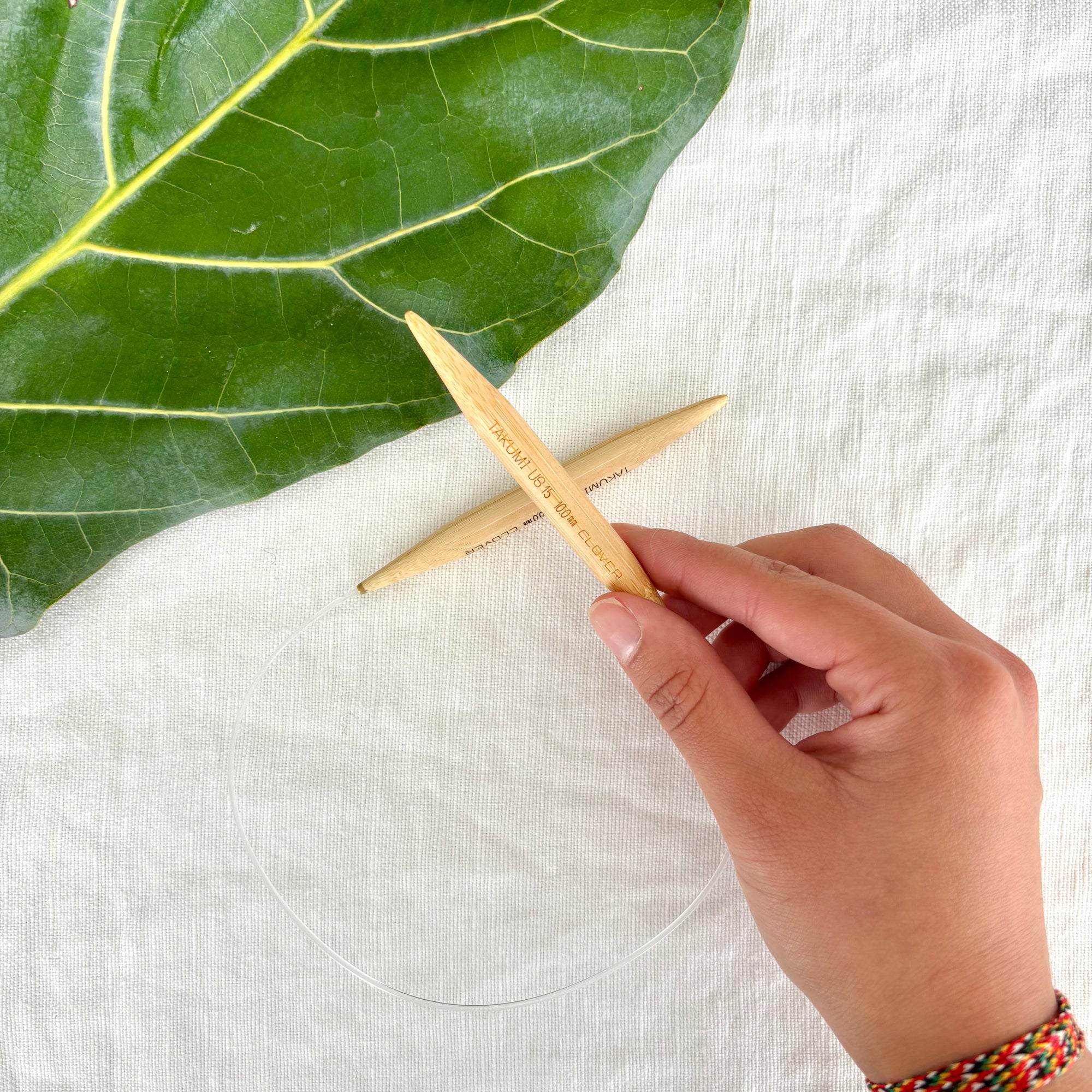 Hand holding wooden knitting needles with a green leaf on a white fabric background
