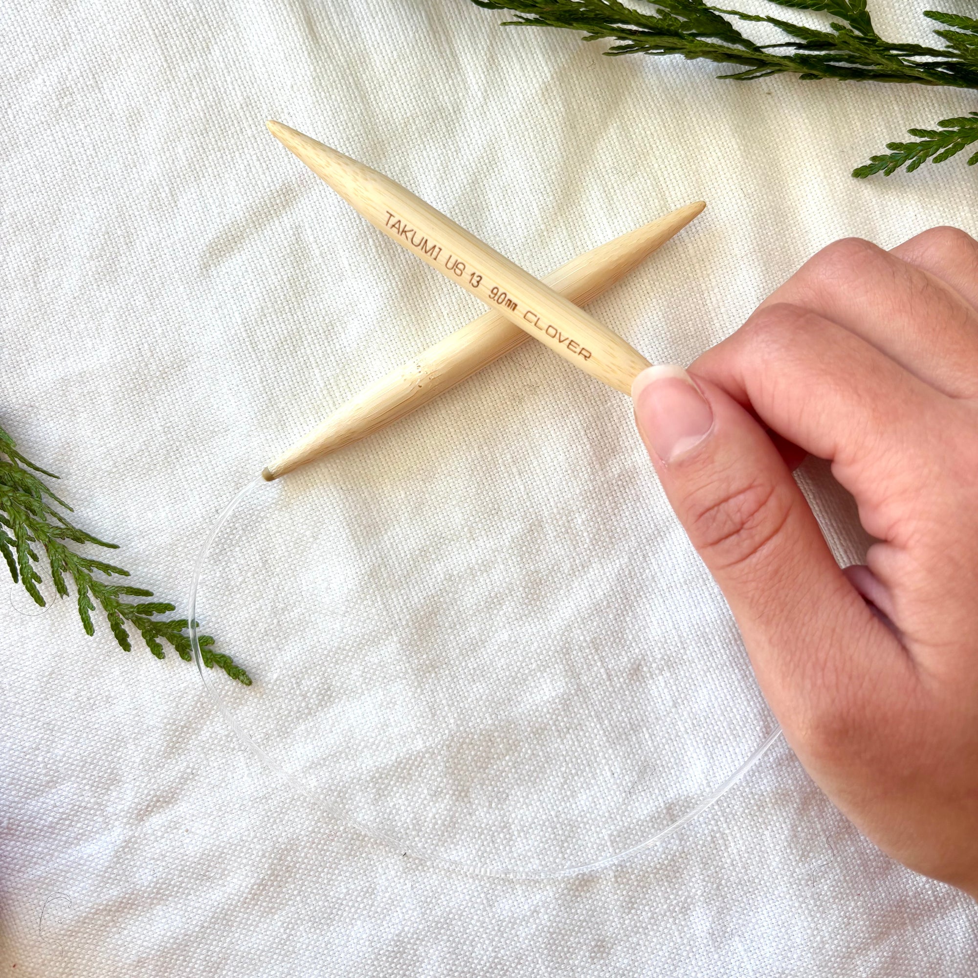 Wooden knitting needles with a hand holding one, on a white fabric background with greenery.