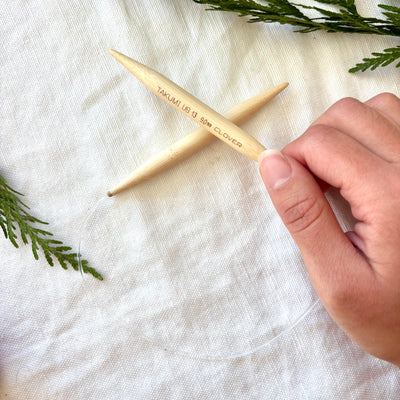 Wooden knitting needles with a hand holding one, on a white fabric background with greenery.